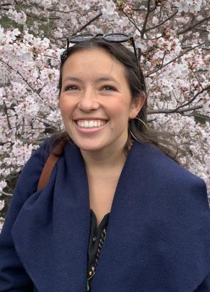 Gabriela Negrete-García, a UCAR Fellow, is wearing a blue jacket as she stands in front of a cherry blossom tree