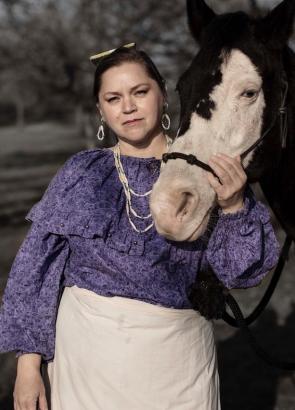 Paulette Blanchard, a UCAR fellow, looks directly into the camera while wearing a purple blouse, holding the nose of a black and white horse. 
