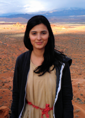 Samar Minallah looks at the camera wearing a beige dress with red belt and black hoodie, while standing in the middle of a red earth desert